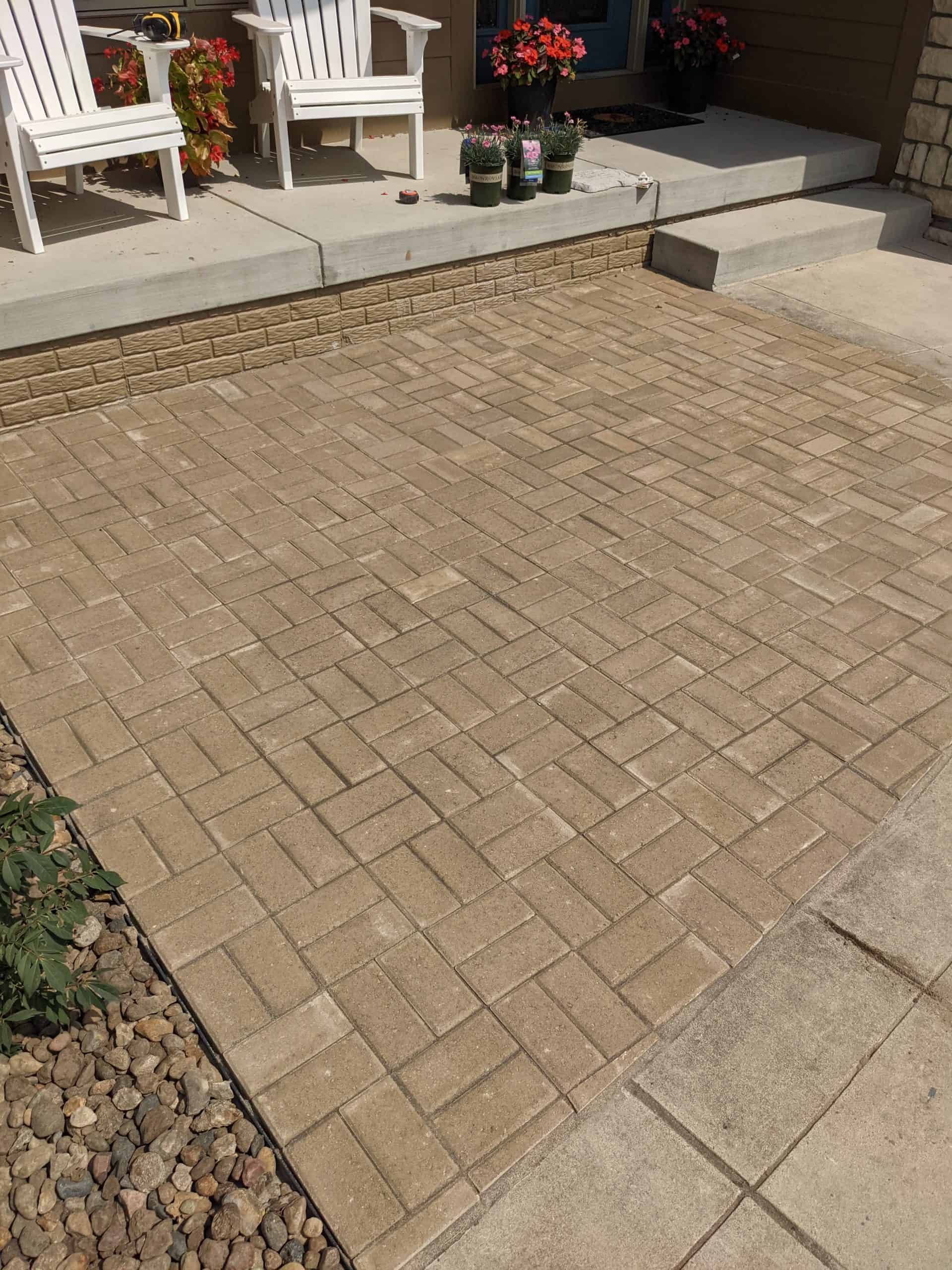 a tan brick patio off the front porch of a house with flower pots and lawn chairs outside